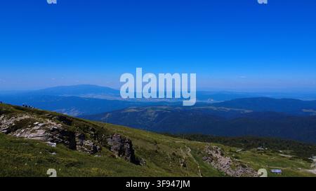 A breathtaking panoramic view from a mountain summit overlooking layered green ridges and distant hazy peaks under a vast clear blue sky. Stock Photo