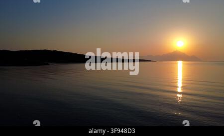 A serene sunset scene with the sun setting behind distant mountains, casting a golden reflection across calm ocean waters. Stock Photo