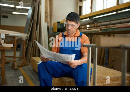 Skilled worker carefully examining detailed blueprints amid tools and timber in quiet workshop Stock Photo