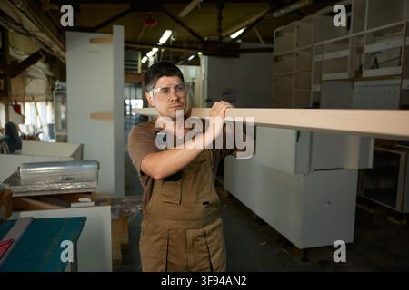 Skilled craftsperson carefully carries raw wood across busy industrial work area with safety gear Stock Photo