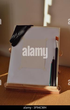 Tabletop wooden artist easel with blank canvas, a black artist glove and pens and brushes Stock Photo