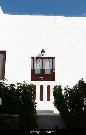 Whitewashed wall with a dark red shuttered window and green shrubs under a blue sky, Mykonos, Greece. Stock Photo