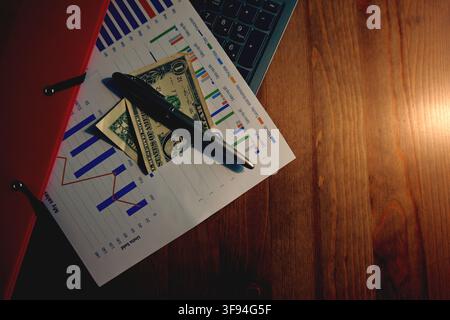 Business documents displaying data charts with a pen and dollar bills placed on a wooden desk, symbolizing financial analysis, investment, and economi Stock Photo