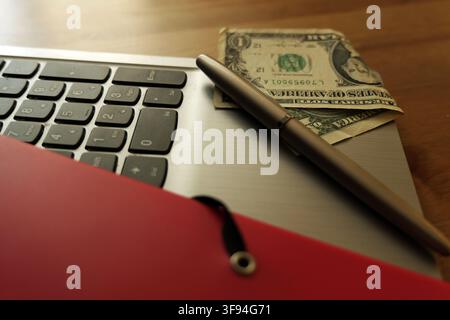 Dollar bill, pen, and red folder resting on a laptop keyboard, representing concepts of online business, digital finance, money management, and modern Stock Photo