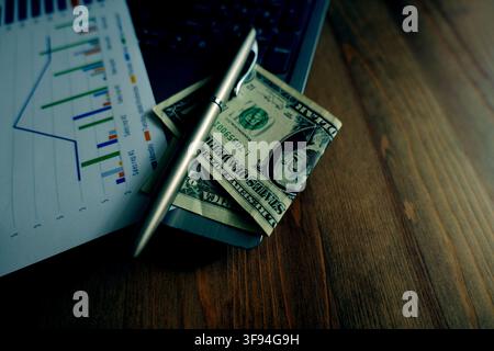 Financial spreadsheet with bar graphs and line charts, a pen, and us dollar bills resting on a dark wooden desk, representing business growth, investm Stock Photo
