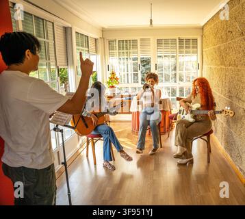 Music rehearsal with young adults playing guitar, bass, trumpet and guiding hand gestures in sunlit home studio Stock Photo