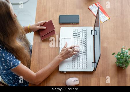 A top view of a woman holding passports while typing on a laptop at a table. Online flight booking, travel planning at home, vacation preparation conc Stock Photo