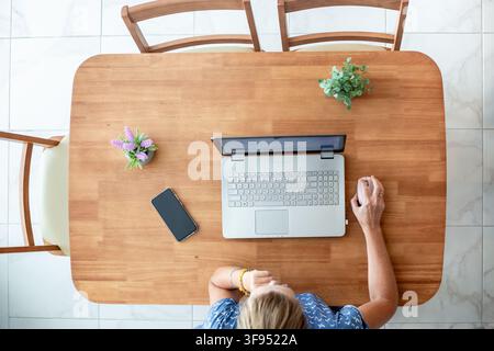 A top down view of a blonde woman sitting at a wooden desk with a laptop. Online remote work, home office freelance, digital workspace layout. Stock Photo