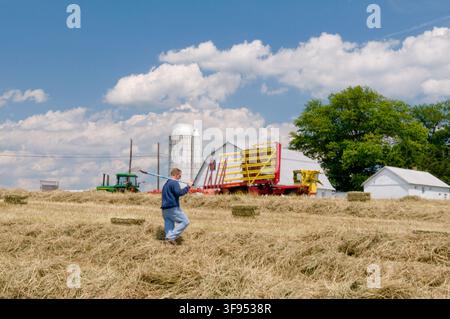 A man stands near a barn, silo, and tractor, surrounded by hay, in an agricultural setting, by Eric Vance on June 20, 2008. Stock Photo