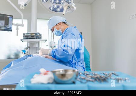 Surgeon in blue scrubs works on patient during a procedure in a hospital operating room. Tools are arranged nearby and lights are on. Stock Photo