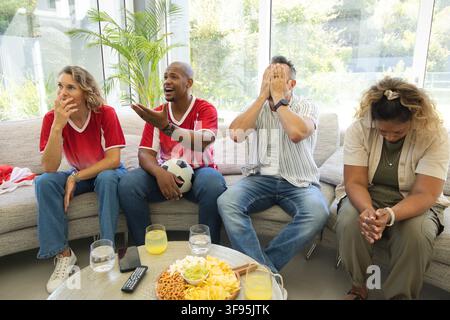 Diverse friends wearing red jerseys watching soccer match at home, holding soccer ball and snacks. Quartet, spectators, livingroom, sofa, daylight, wi Stock Photo