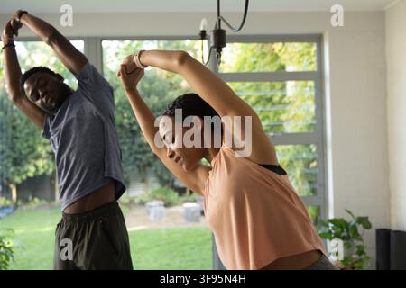 African American couple doing side stretches in living area near glass doors wearing wrist tracker. Duo, home, fitness, wellness, stretching, backyard Stock Photo