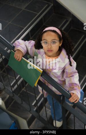 African American child standing on tiled stairwell grasping metal handrail holding green folder Stock Photo