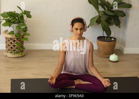 Woman sitting crosslegged meditating on black mat at home wearing activewear, headphones, diffuser Stock Photo