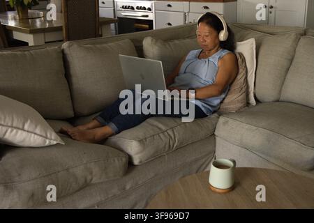 Senior Asian woman reclining on gray sofa at home wearing headphones typing on laptop, copy space Stock Photo