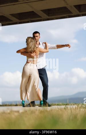 Couple dancing outdoors under a concrete overhang, practicing partner dance in a close embrace with flowing skirt and scenic sky backdrop Stock Photo