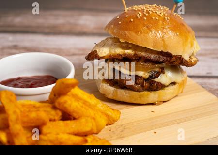 Double hamburger with fried egg, melted cheese and caramelized onions, presented on a wooden board with French fries and ketchup. Stock Photo