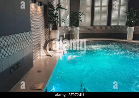 uxurious and modern indoor hydrotherapy pool featuring sparkling turquoise water, a cascading water jet feature, decorative patterned tile walls, and Stock Photo