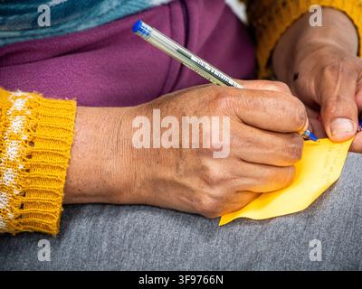 Close-up photograph of a person writing handwritten notes on a yellow paper with a blue ballpoint pen. The image focuses on hands, handwriting, concen Stock Photo