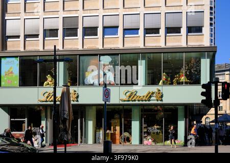 A festive storefront window display in Frankfurt, Germany, showcases Lindt's chocolate making demonstration. The scene is set on a sunny city street w Stock Photo