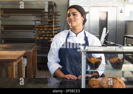 Female baker working behind counter wearing chef coat and apron with glass display of muffins Stock Photo