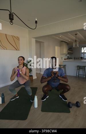 African American couple doing squats on green mats at home in sportswear holding weights. Gym, fitness, training, duo, partners, dumbbell, kettlebell Stock Photo