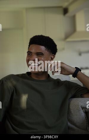 Adult African American man laughing, sitting on light sofa at home, wearing olive tee, black watch. Subject, couch, livingroom, kitchen, candid, lifes Stock Photo