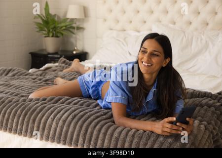 Indian woman lying on king-size bed holding dark smartphone, wearing blue satin pajamas, smiling Stock Photo