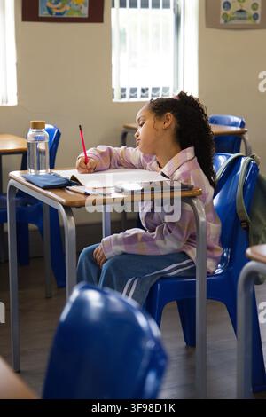 African American schoolgirl in purple jacket writing on paper at desk in classroom with red pencil. Student, table, blue, tablet, water, backpack, day Stock Photo