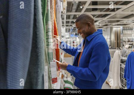 A young African man in a blue jacket examines colorful fabric swatches in a modern retail store. He appears focused and engaged in shopping Stock Photo