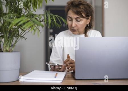 Portrait serious pensive mature business woman in white clothes thinking or making decision, using laptop computer at home, holding pen, brainstorming Stock Photo
