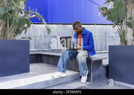 A young Black man sits on concrete steps, wearing a blue jacket and orange shirt. He holds a smartphone and looks thoughtfully into the distance Stock Photo
