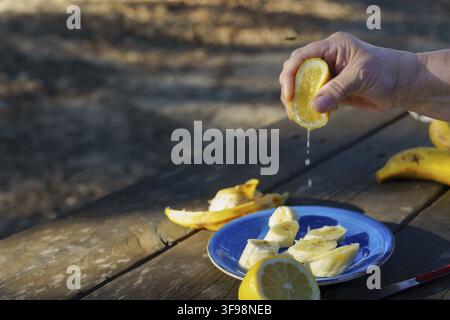 Hand squeezing fresh lemon juice over sliced bananas on a blue plate, preparing a healthy snack on a wooden table outdoors Stock Photo