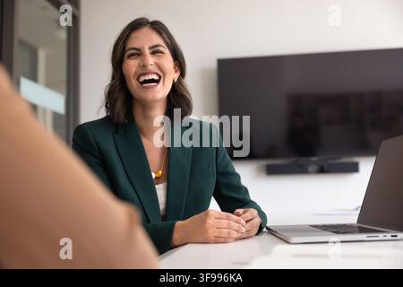 Latina businesslady in elegant jacket sit at office desk with laptop, laughing while talking to female colleague or client engaged in positive communi Stock Photo