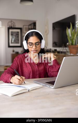 Indian woman in headphones sits at home with laptop and notebook, focused on taking notes during online class or remote meeting, writing down importan Stock Photo