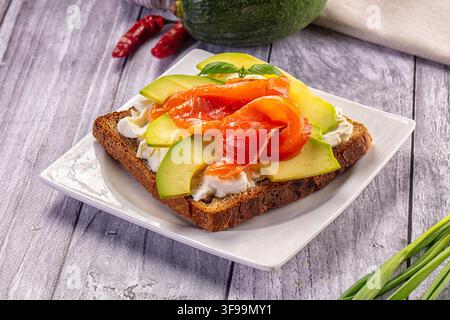 Smoked salmon avocado slices layered over cream cheese on whole grain toast. Served on white plate with basil garnish. Rustic wooden background with r Stock Photo