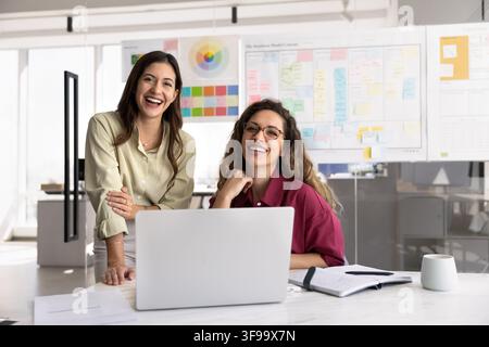 Two female designers working together at desk with laptop, smiling at camera, reviewing creative project, posing for professional corporate office por Stock Photo