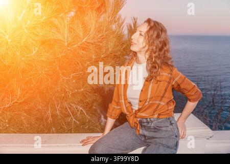 Sunset woman balcony sits on a white terrace overlooking the sea during golden hour with warm light through pine trees Stock Photo