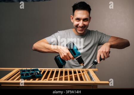 Man smiling looking at camera broadly using cordless drill to assemble wooden furniture, playful diy moment showing imperfect handiwork, humor and Stock Photo