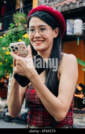 A smiling young woman wearing a red beret and glasses holds smartphones while walking through a busy street. Stock Photo