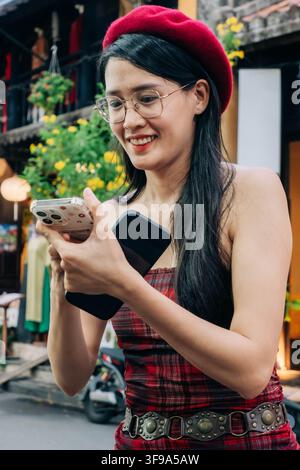 A young woman in a red beret and glasses looks at her smartphone among colorful street surroundings. Stock Photo