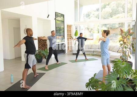 Diverse group practicing Warrior II on yoga mats in living room in sportswear with water bottle. Fitness, instructor, practice, home, modern, bright, Stock Photo