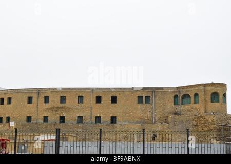 A wide shot featuring a long, curved yellow-stone masonry building with small rectangular shuttered windows, arched top openings Stock Photo