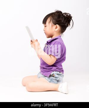 Toddler girl sitting on floor and using digital tablet, focused child learning online or watching educational video, white background. Stock Photo