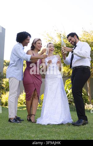 Diverse wedding group on grassy lawn beside wall, bride wearing white gown, groom pouring wine Stock Photo