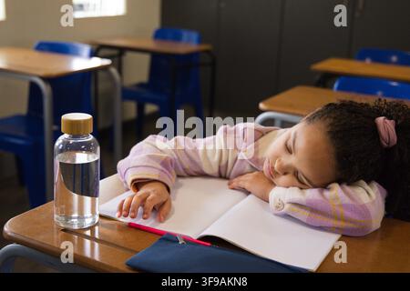 Female child in classroom resting on arms over notebook at wooden desk, reaching for water bottle Stock Photo