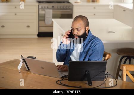 Bearded man talking on smartphone and typing on laptop at home kitchen table with monitor Stock Photo
