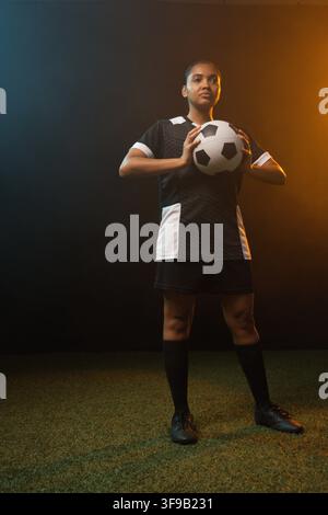 Adult female soccer player standing on synthetic-turf holding soccer ball in kit, rim-lit two-tone Stock Photo