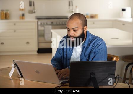 African American man working at kitchen table using silver laptop and black monitor, wearing denim Stock Photo
