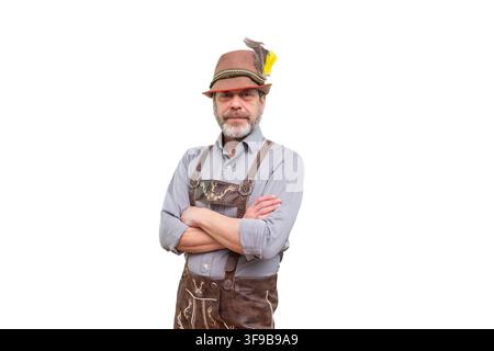 A bearded middle aged man in traditional Bavarian clothing stands with crossed arms against a light isolated background. Stock Photo
